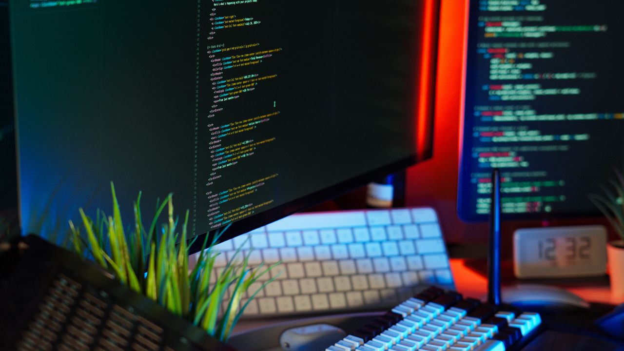 About A vibrant workspace showing computer monitors with code, keyboard, and tech accessories.