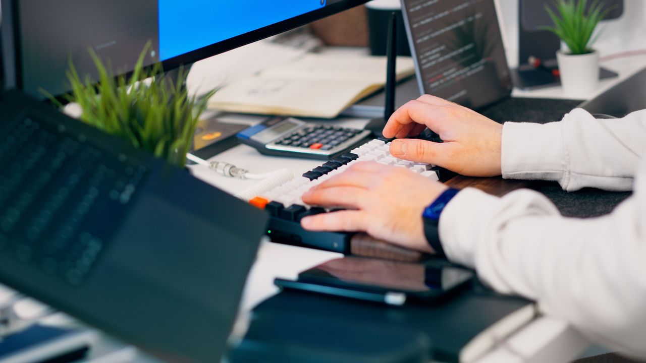Services A close-up of a person typing on a keyboard in a modern tech workspace with gadgets and a monitor.
