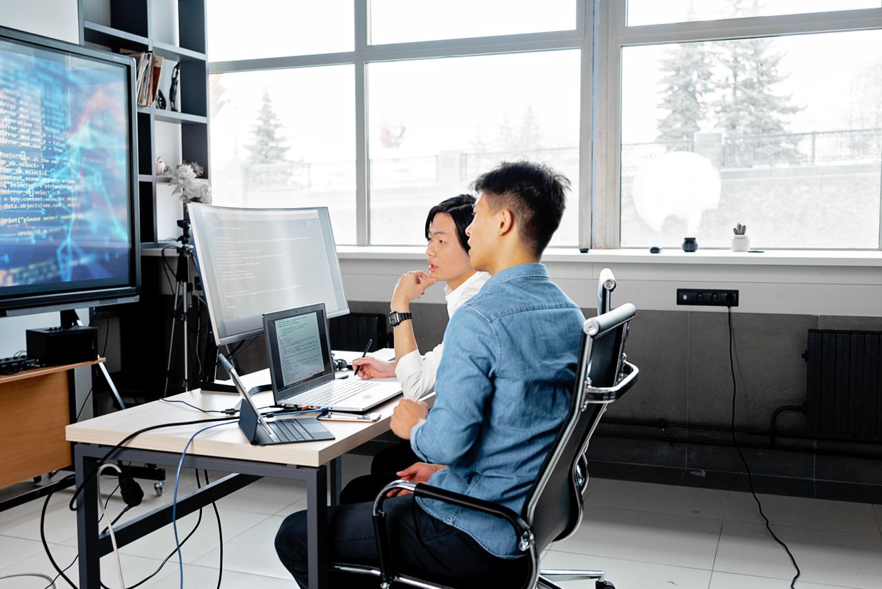 Services Two men analyzing code on computers in a modern office setting.